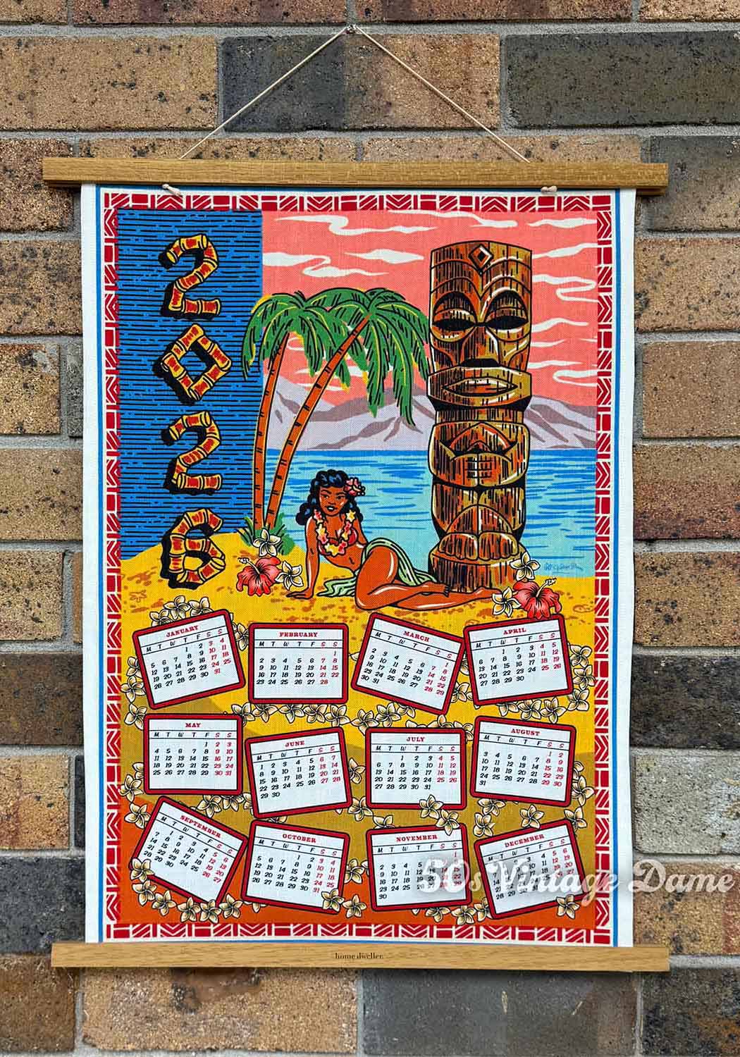 2026 Tiki Calendar teatowel with tropical design featuring a tiki statue and palm tree, hanging on a brick wall.