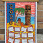 2026 Tiki Calendar teatowel with tropical design featuring a tiki statue and palm tree, hanging on a brick wall.