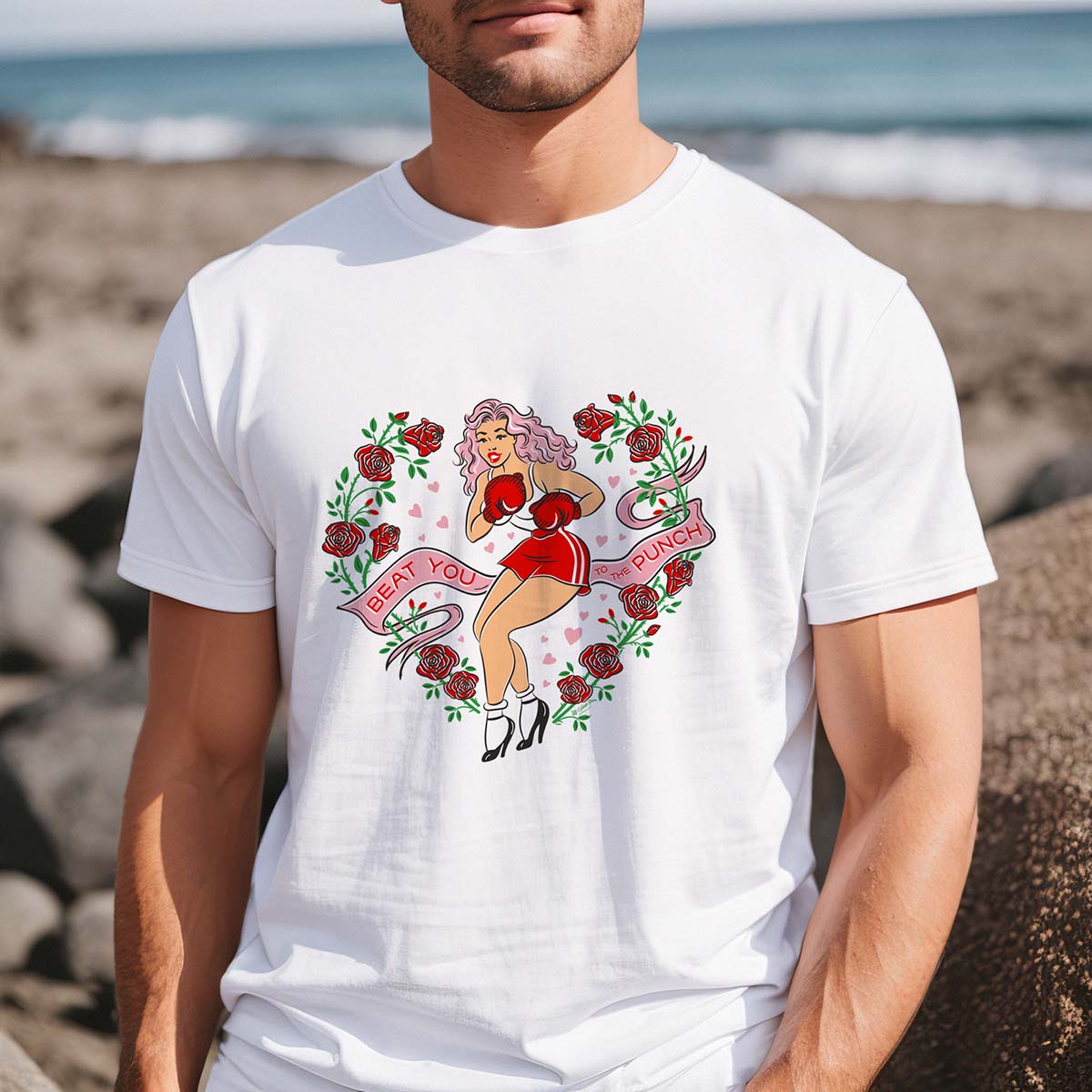 Person wearing a white t-shirt with a graphic design of a boxing pinup lady on a beach.