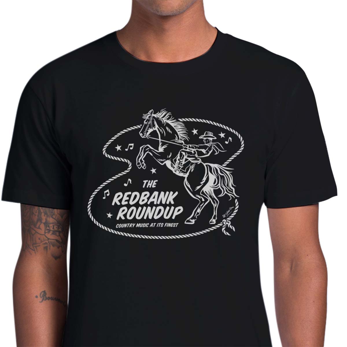 Black unisex t-shirt with a cowboy riding a bucking bronco. Text says "The Red Bank Roundup - country music at its finest" A lasso, stars and music notes surround the design.