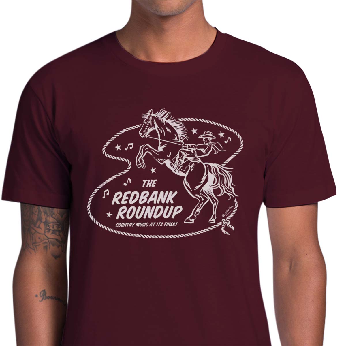 Burgundy unisex t-shirt with a cowboy riding a bucking bronco. Text says "The Red Bank Roundup - country music at its finest" A lasso, stars and music notes surround the design.