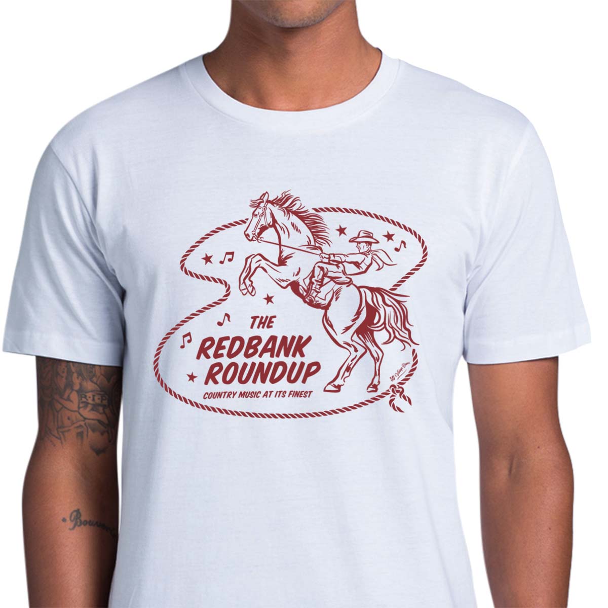 White unisex t-shirt with a cowboy riding a bucking bronco. Text says "The Red Bank Roundup - country music at its finest" A lasso, stars and music notes surround the design.