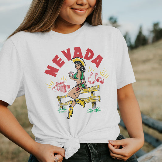 Vintage-style illustration by 50s Vintage Dame of a pinup cowgirl sitting on a wooden fence holding a Royal Flush. Headline says "Nevada" . White short-sleeved tee shirt.